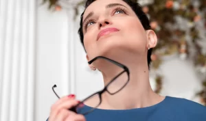 Image of a young woman with short black hair holding her glasses in her hands and anxiously looking up as if she is contemplating something. This image represents the anxiety and uncertainty one may feel before a special occasion and how Kybella can help boost confidence and provide a more defined and balanced appearance.