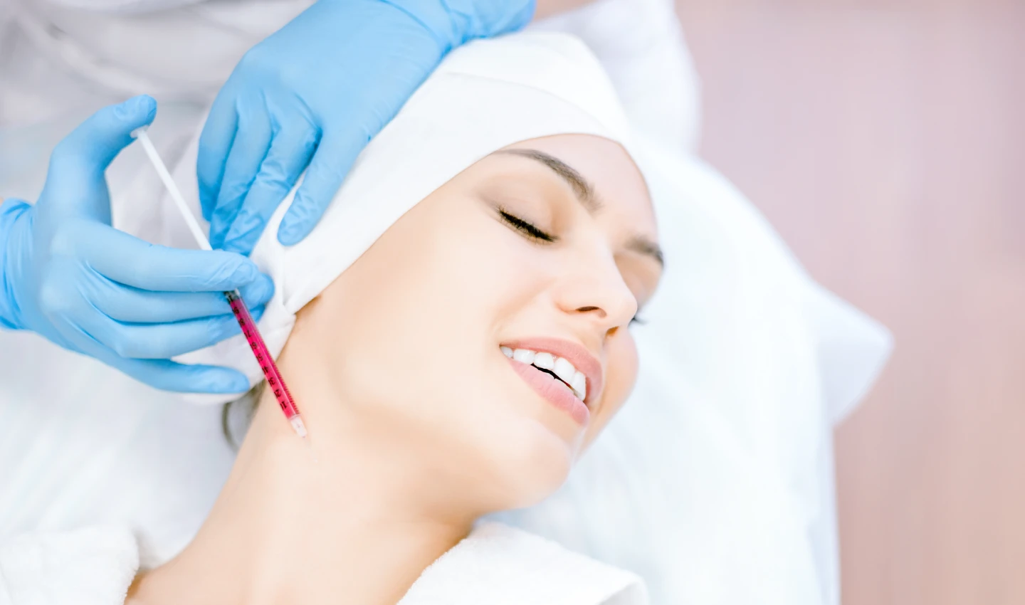 A woman receiving dermal filler treatment for postpartum facial rejuvenation, smiling happily during the procedure.
