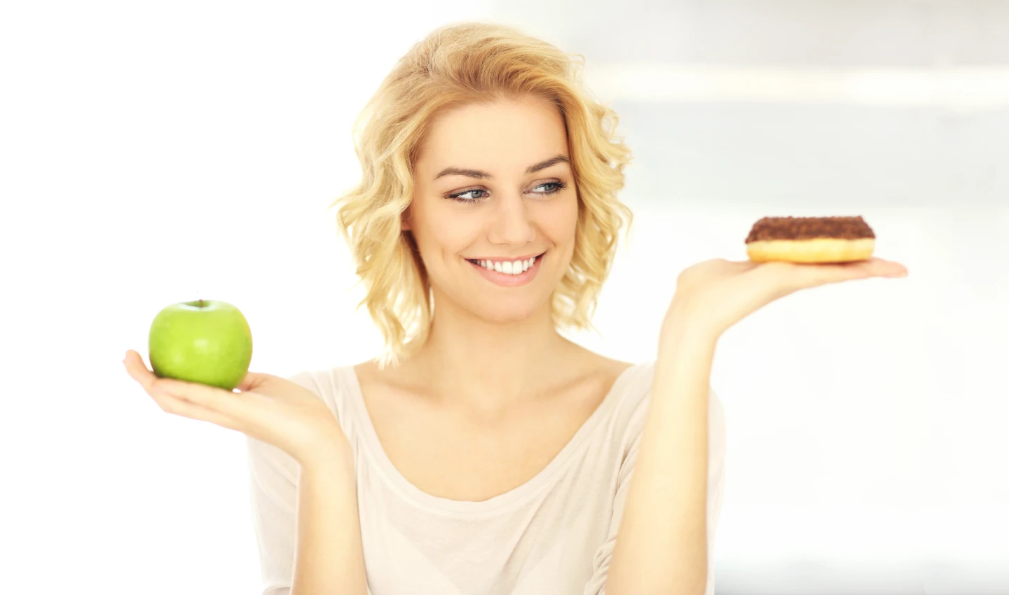 Woman with a smile holding an apple and a chocolate donut, reflecting her healthy lifestyle after teeth beauty surgery.