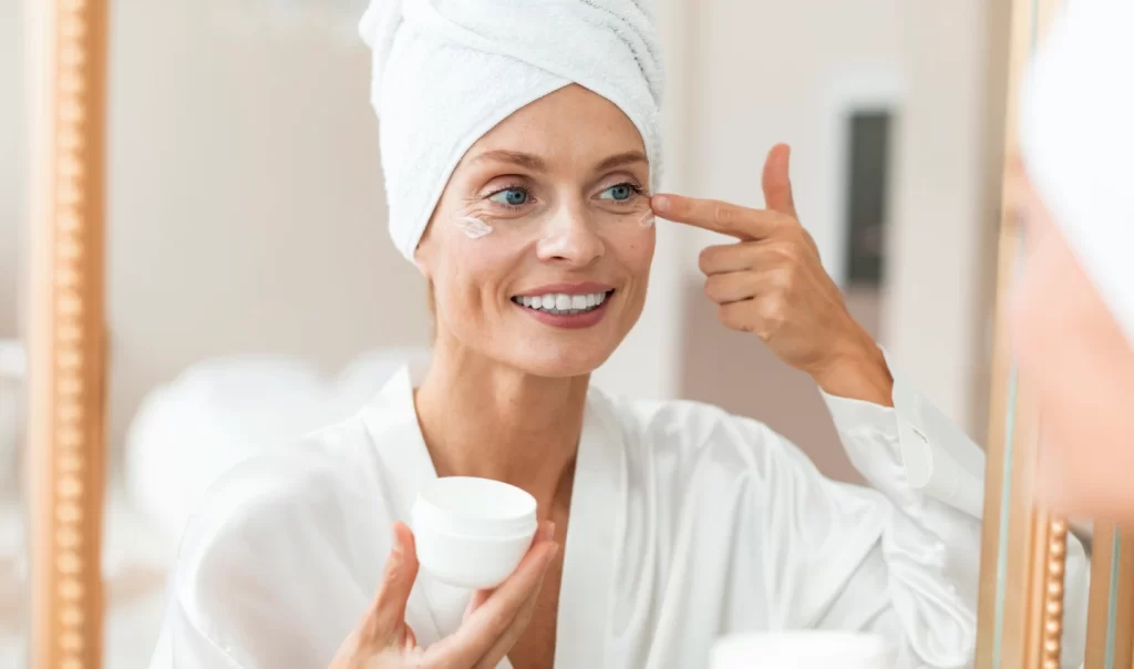 A mature woman smiles while applying eye cream to her under-eye area, enjoying the many benefits it provides for her skincare routine, including reducing fine lines, wrinkles, and dark circles.