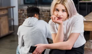 A woman sits upset while a man stands with his back turned in a conversation about coping with societal pressure and facelift surgery expectations.