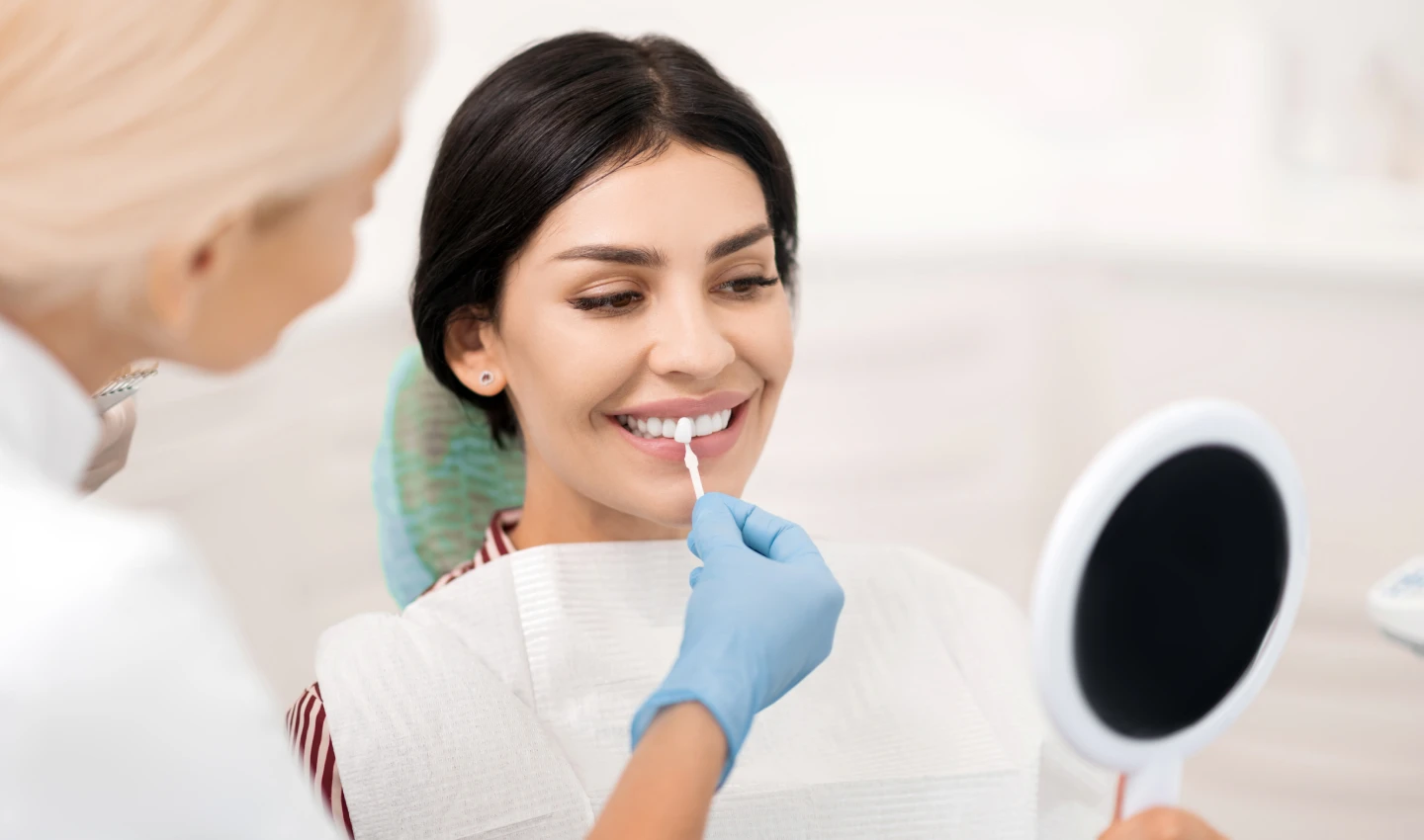 A patient smiles while a dentist performs dental bonding on her teeth. Dental bonding benefits include non-invasiveness, immediate results, customization, natural-looking appearance, affordability, and long-lasting effects.