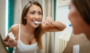 Woman brushing teeth and smiling to showcase the results of proper post-operative care after cosmetic dental surgery.