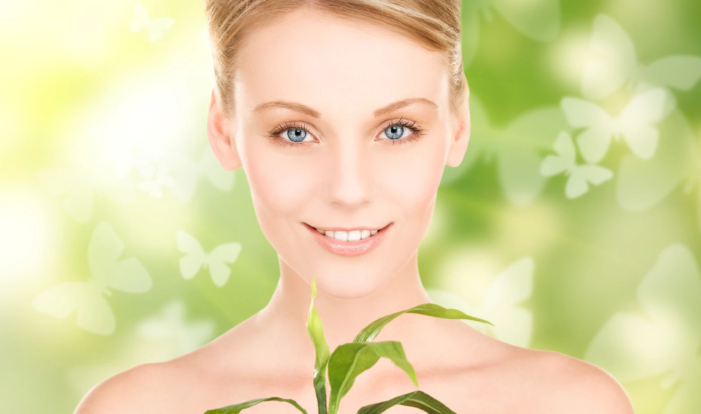 A young woman with a glowing complexion smiles at the camera against a backdrop of plants and greenery.