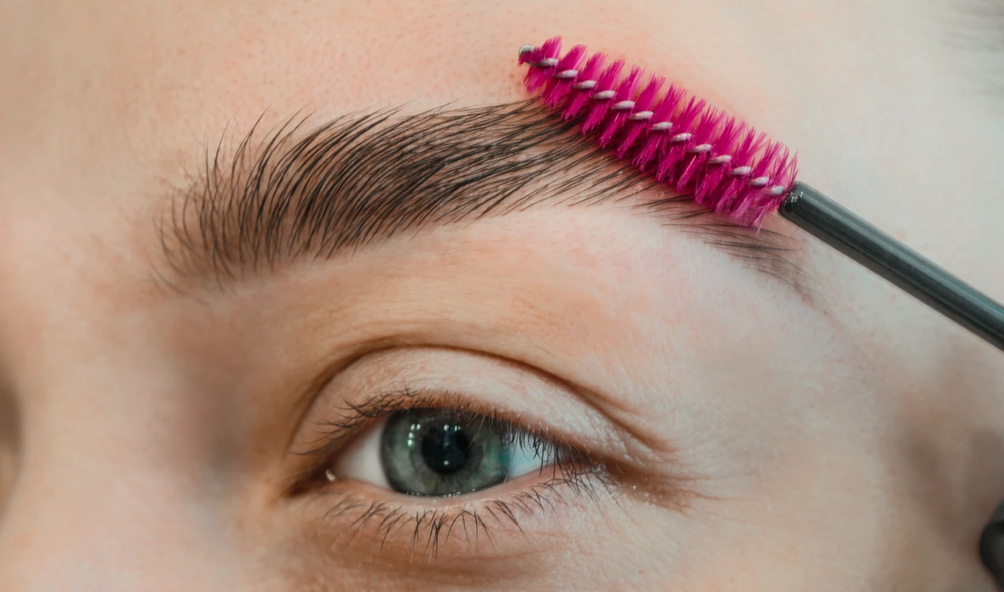 A pink spoolie brush being used to shape and define the brown eyebrows of a young woman, highlighting the importance of prepping your brows before filling them in.