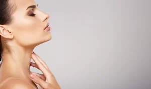 Image of a young woman with closed eyes, touching her neck and chin with her long, beautiful fingers. The image represents the natural beauty enhancements that can be achieved with Kybella, a non-surgical injectable treatment for reducing submental fat and enhancing the facial profile.