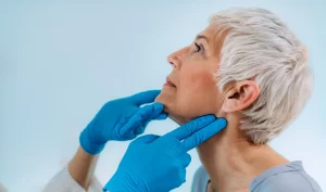 Image of a dermatologist wearing blue medical gloves examining the chin of a middle-aged woman with gray hair who is smiling. The image represents the process of receiving Kybella treatment from a qualified dermatologist to achieve a youthful and defined neck.