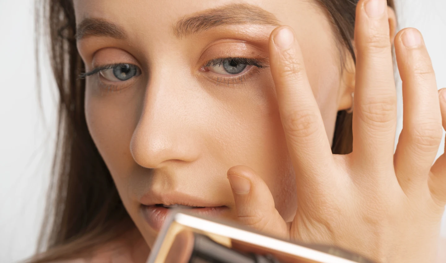 A woman with glowing skin applying a nude-colored eye shadow with her index finger above her eyes, showcasing the ease and simplicity of using neutral eye shadow palettes to create an effortlessly elegant eye makeup look.