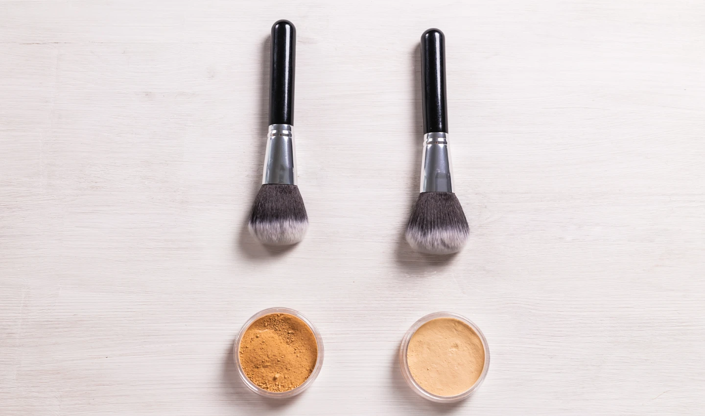 Two brushes of foundation, one on top of two batches of liquid foundation and the other on top of two batches of powder foundation, emphasizing the differences between the two types of foundation.
