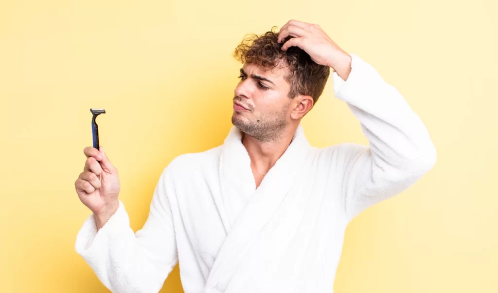 Man holds shaving supplies, including a razor and shaving cream, in his hands in front of a bathroom mirror, preventing razor burn.