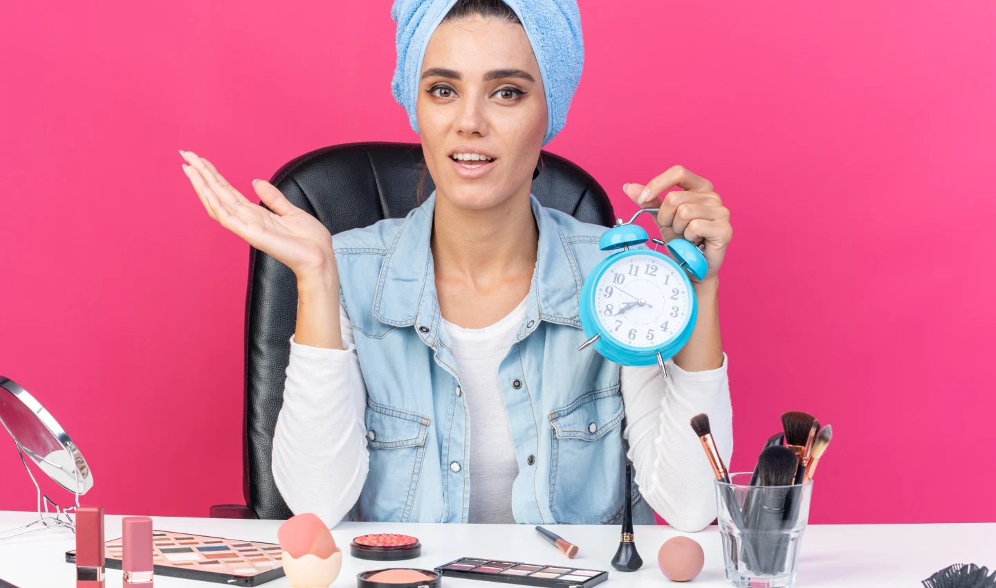 A young woman in sky blue jeans and coat is holding a clock in her hand while looking at the camera, highlighting the importance of making your foundation last all day with these long-lasting hacks.
