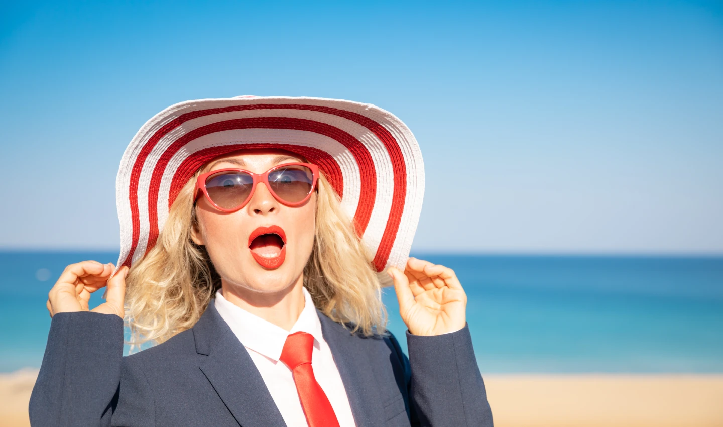Woman with sunglasses standing on a beach, looking at the sun, with a glowing complexion achieved through proper skincare routine.