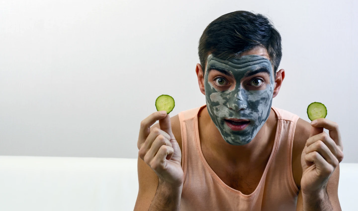 A man wearing a face mask and holding out a cucumber slice, representing a DIY face mask for men's skin care.