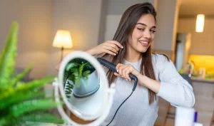 Smiling woman using a hair straightener to protect her hair at home Caption: A woman with a smile on her face straightens her hair using a hair straightener, taking steps to protect her hair.