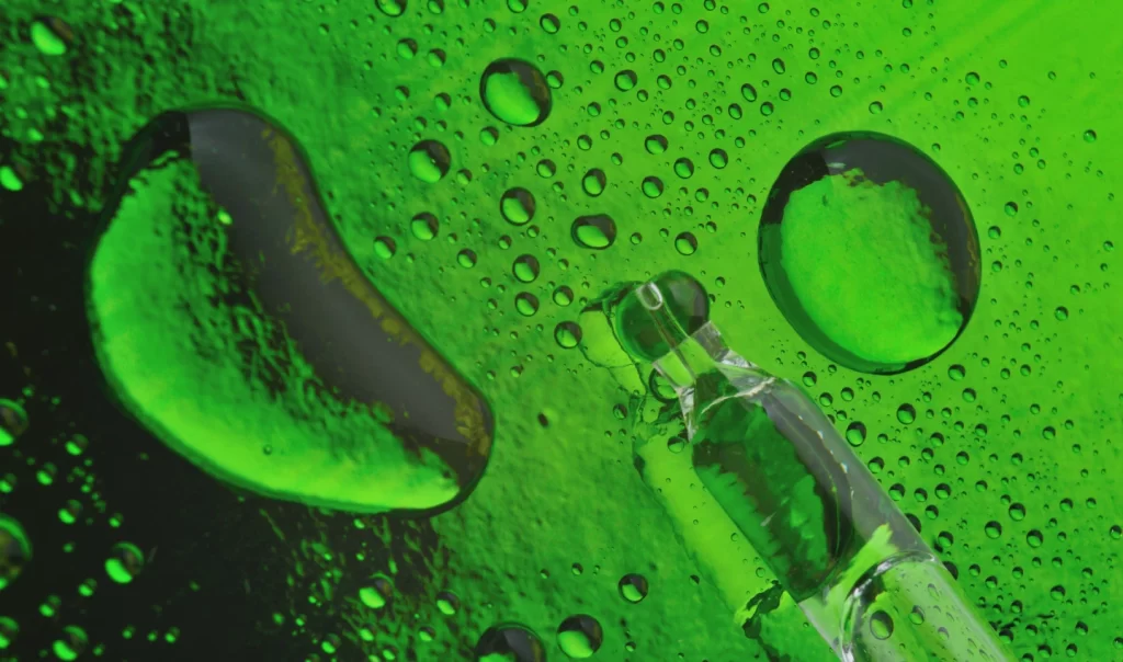 Close-up photo of a cosmetic pipette filled with Alpha Hydroxy Acids, placed on a green background covered in droplets.