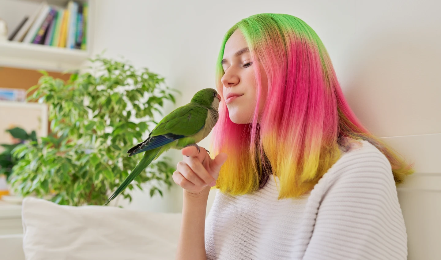 A teenager girl with pastel-colored hair kissing a green quaker parrot up close.