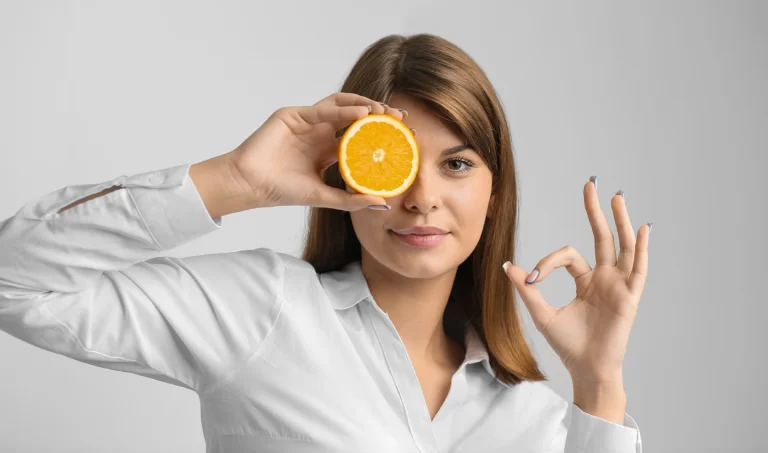 Woman holding a slice of orange over her eye to highlight the benefits of vitamin C for dark skin.