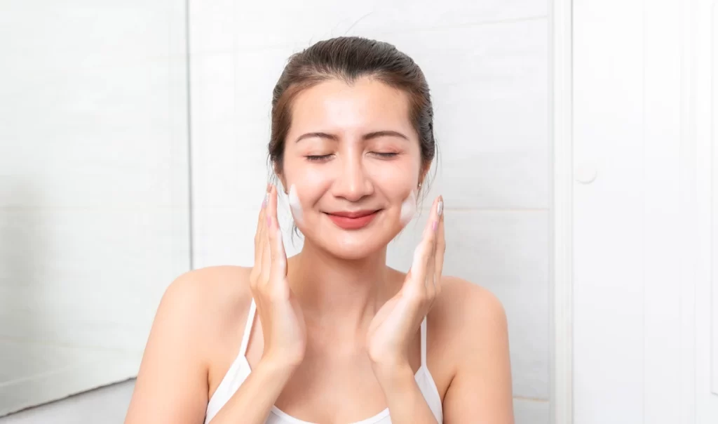 A smiling woman happily applies moisturizers to her face, aiming for even skin tone